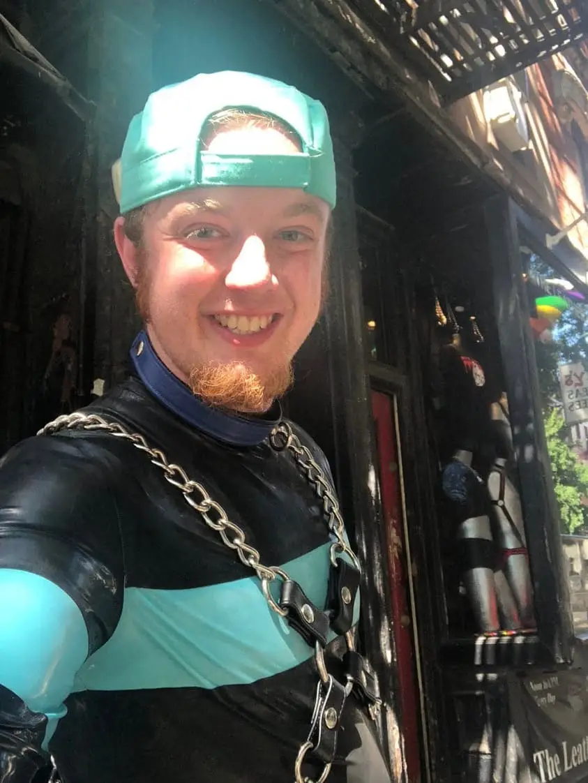 Photograph identified as Luke Wenke smiling outdoors while wearing a backward teal cap, black-and-teal latex clothing, and a metal chain harness.